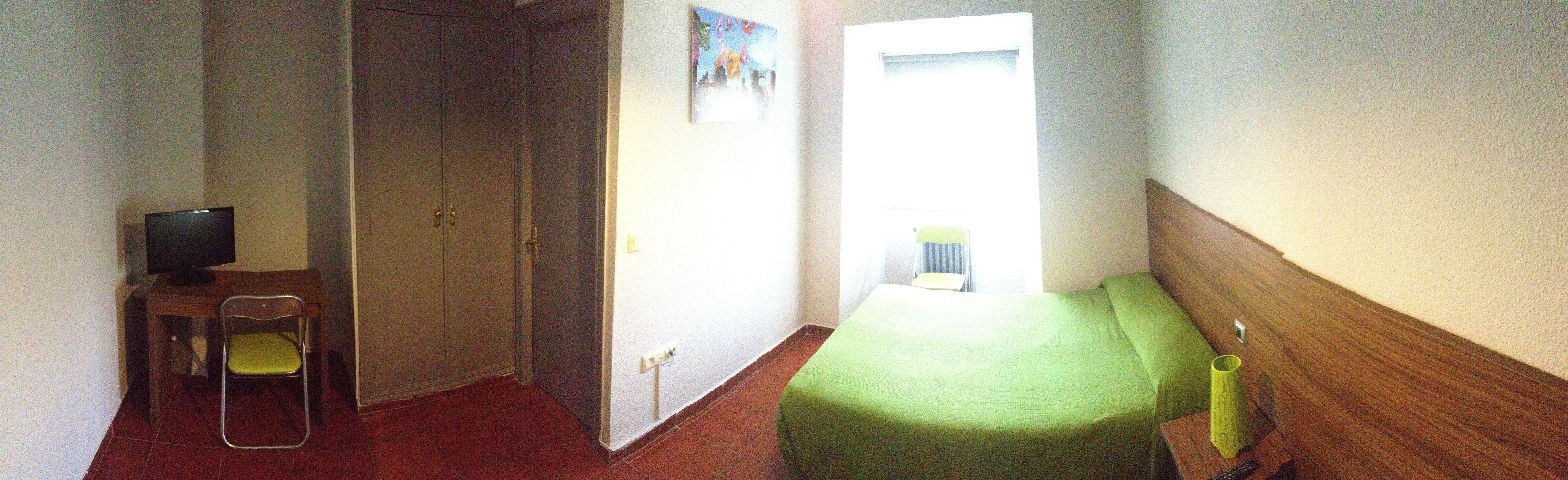 Una habitación pequeña con cama, una silla, televisor y ventana.