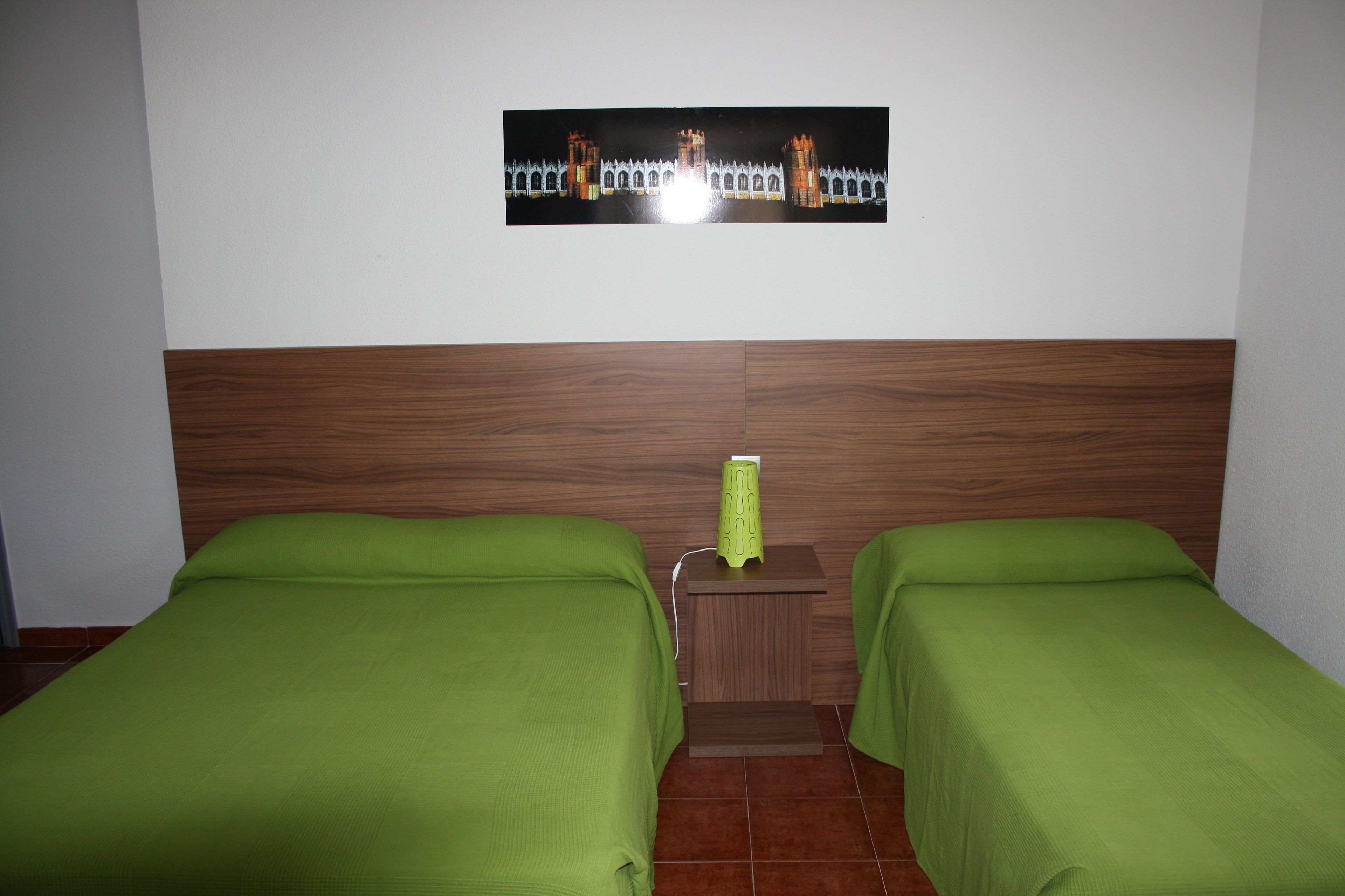 Habitación de hotel con dos camas individuales, cabecero de madera, mesita de noche con lámpara y un cuadro colgado en la pared.