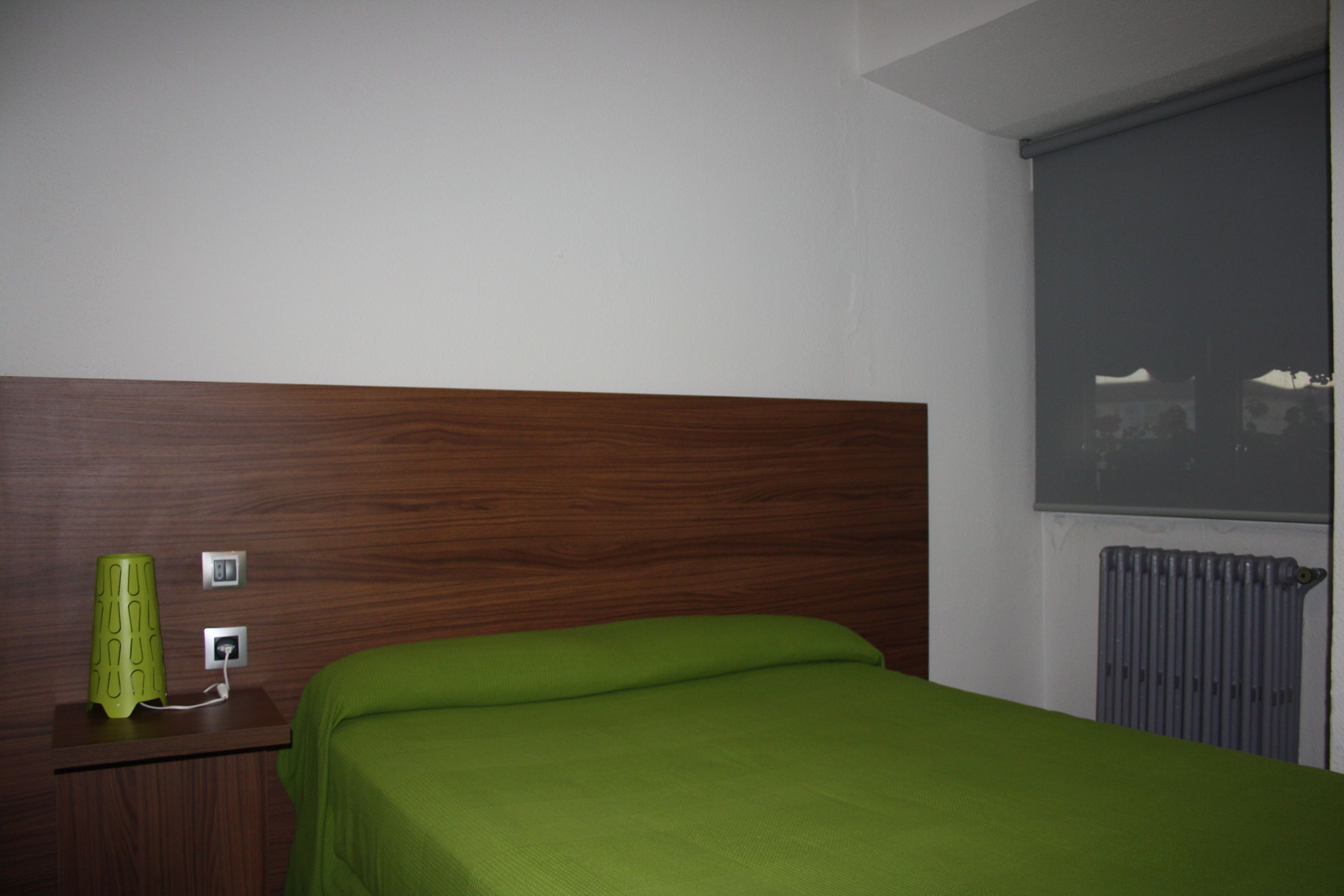 A hotel room with a wooden headboard, a green bedspread, a nightstand with a green lamp, and a window with a view of a building.