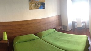 A hotel room with a wooden headboard, a green bedspread, a small bedside table with a lamp, and a painting on the wall.
