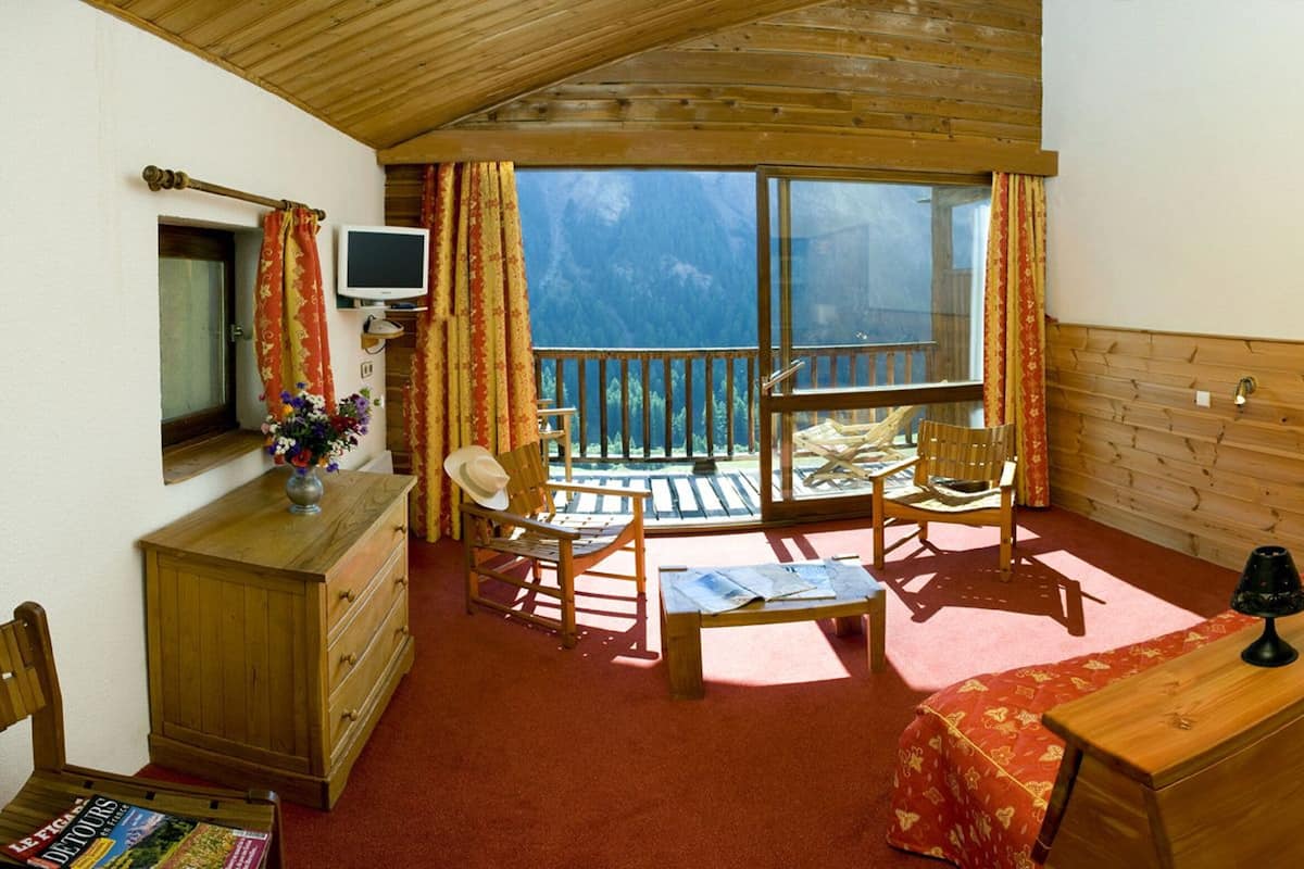 Ein gemütliches Zimmer mit Holzboden, einem Balkon mit Bergblick, einem hölzernen Schreibtisch mit Lampe und einem Stuhl.