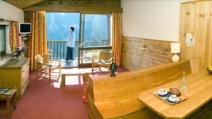 A room with a wooden reception desk, a dining area with a table and chairs, a balcony with a person standing, and a view of the outdoors. - Les Chalets du Villard (Saint-Veran)