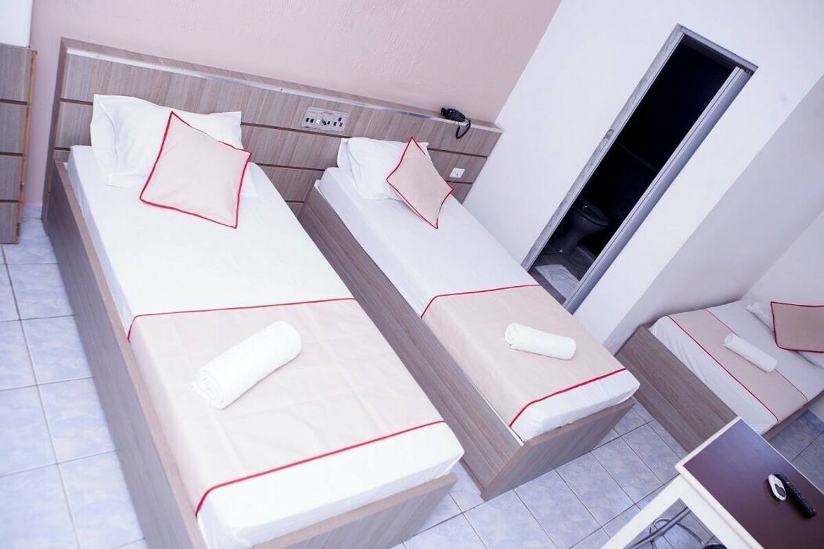 A hotel room with three single beds, each with a red-lined headboard, white bedding, and a rolled towel. There is a small desk with a computer and a phone.