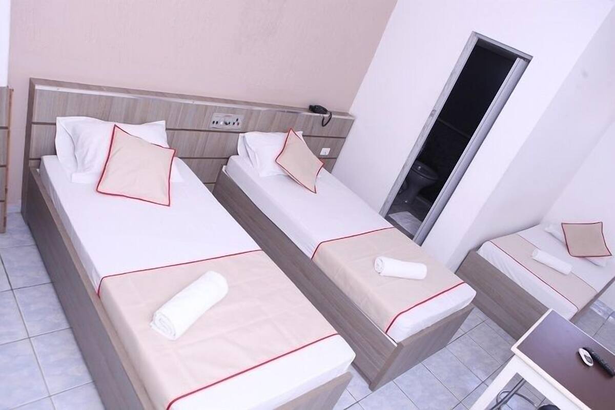 A hotel room with two single beds, each with a pillow and a rolled towel. There is a bedside table with a phone and a small lamp.