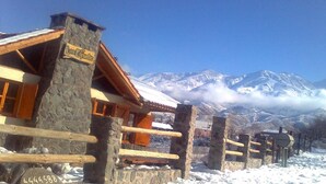 Un chalet de montagne rustique aux murs de pierre et aux poutres en bois, situé dans un cadre spectaculaire avec des montagnes enneigées en toile de fond et un ciel dégagé.