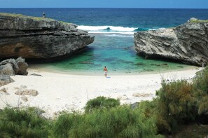 A beach with clear turquoise water, a person standing on the sand, and rocky cliffs.