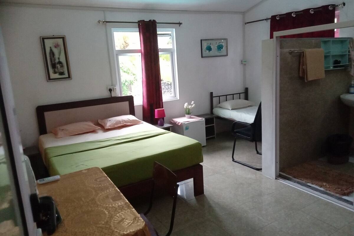 A bedroom with a bed, a desk, a chair, a small table, a window with curtains, and a bathroom visible through an open door.