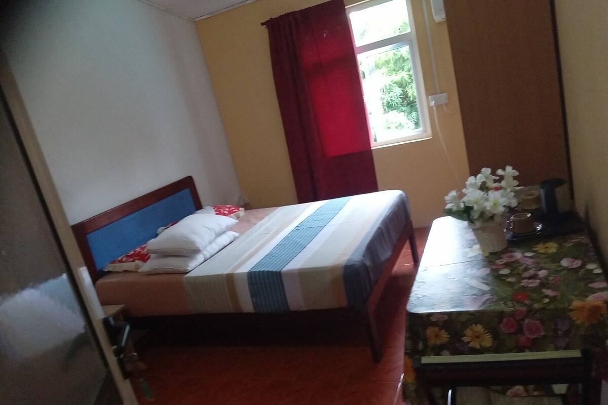A small, neatly arranged bedroom with a bed, a table with a floral tablecloth, and a window with red curtains.