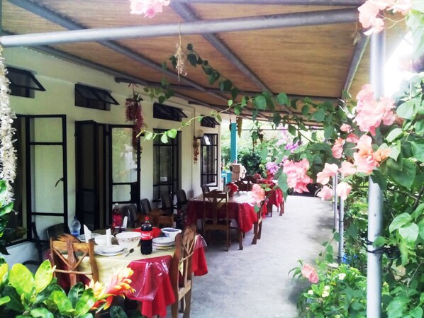 An outdoor dining area with tables covered in red tablecloths, surrounded by lush greenery and pink flowers.
