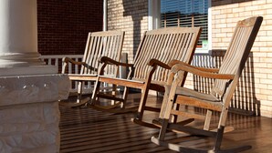 Trois chaises à bascule en bois sont placées sur une terrasse en bois, avec un mur de briques et une fenêtre en arrière-plan.