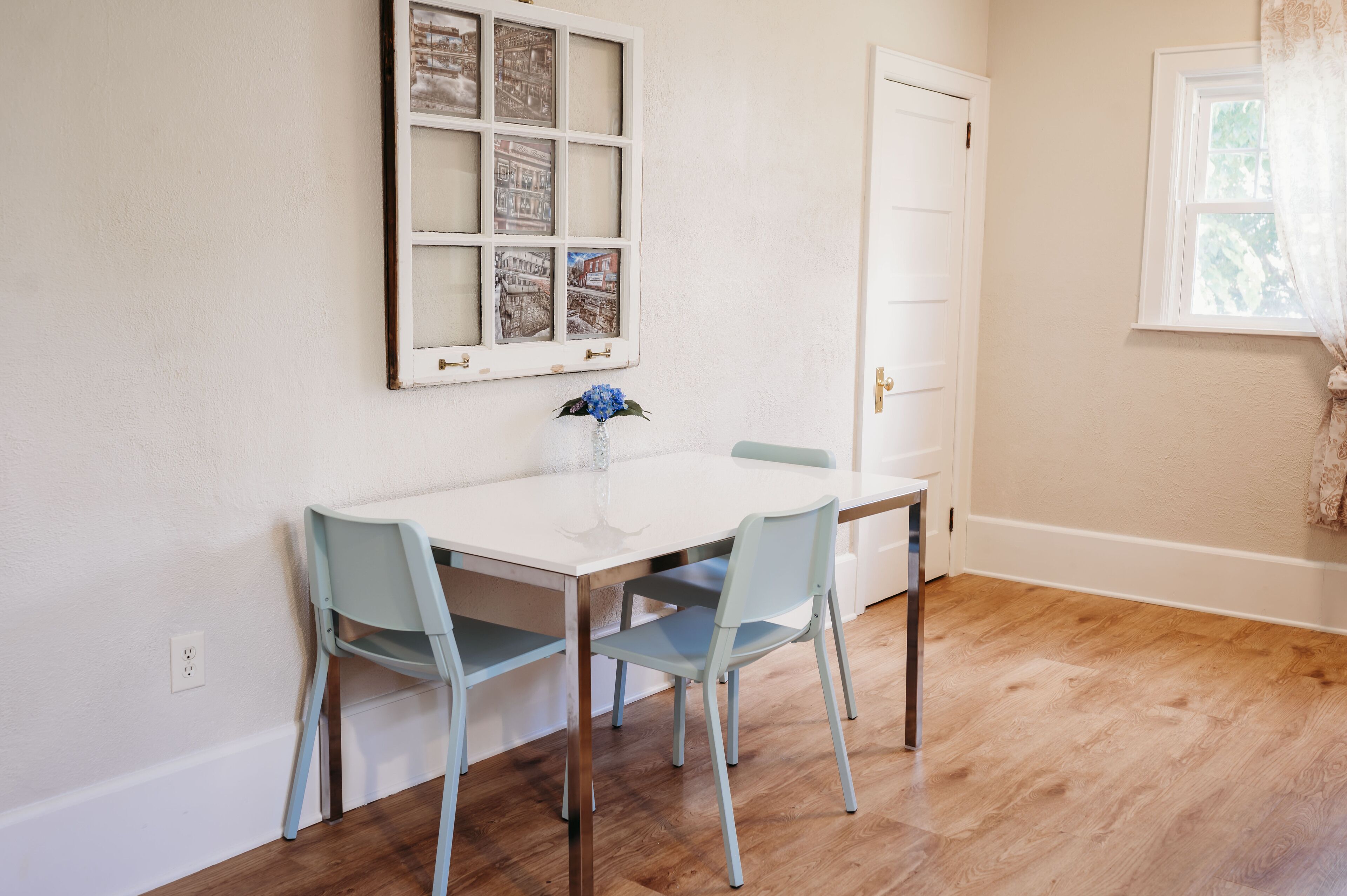 A dining area with a white table and two light blue chairs, a window with a curtain, and a door with a white frame.