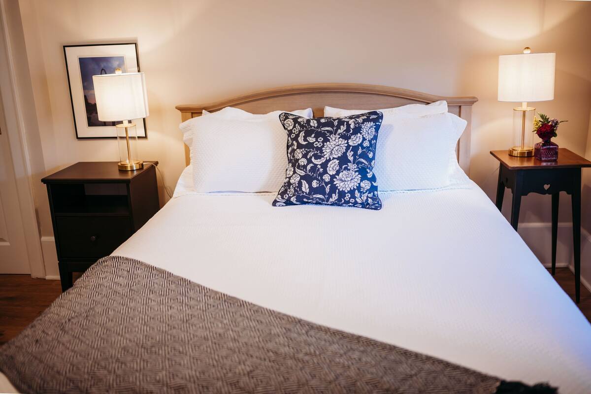 Una cama bien tendida con sábanas blancas y una almohada azul con estampado floral, flanqueada por dos mesitas de noche con lámparas y un cuadro en la pared.