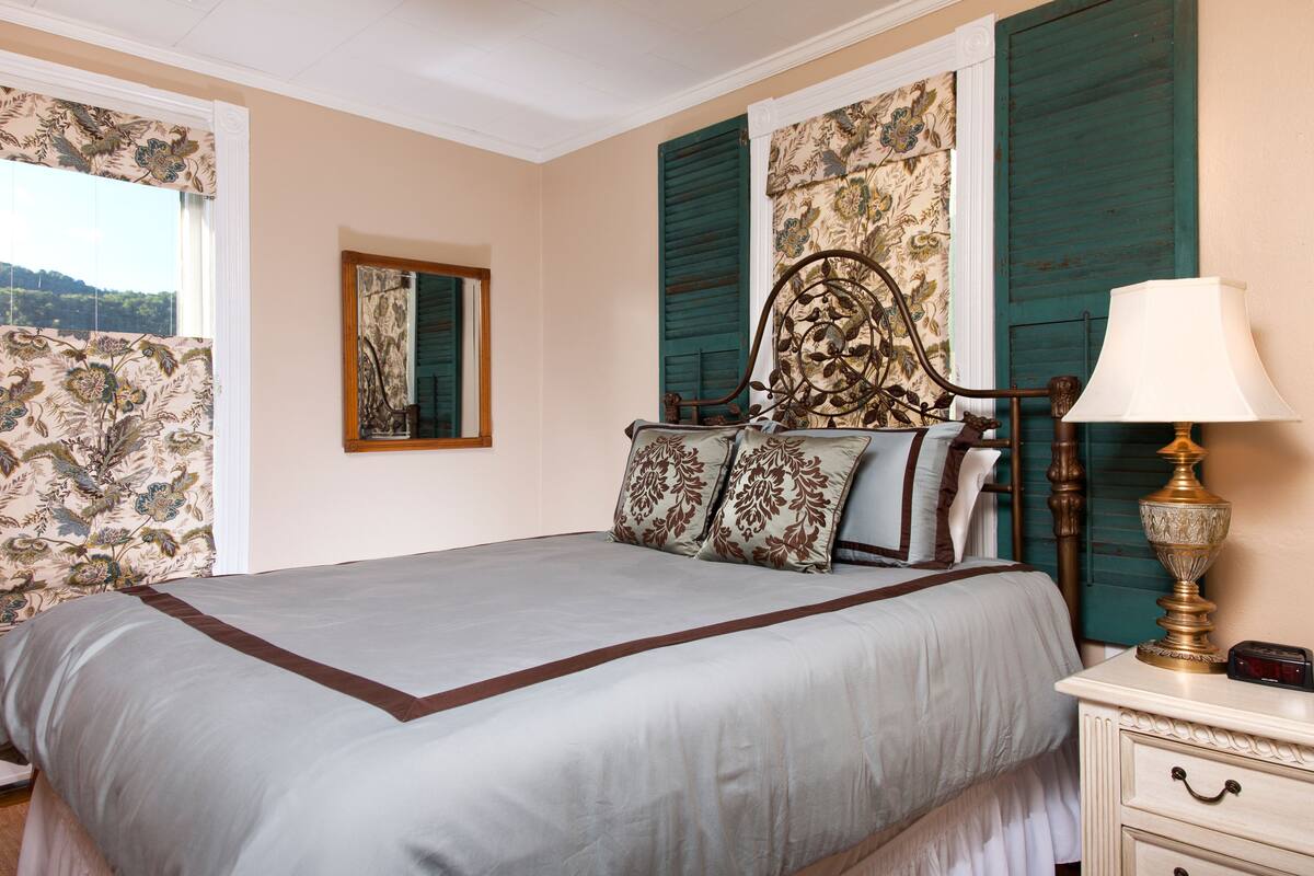 Una cama bien hecha con cabecera floral, una mesita de noche con lámpara y una ventana con contraventanas.