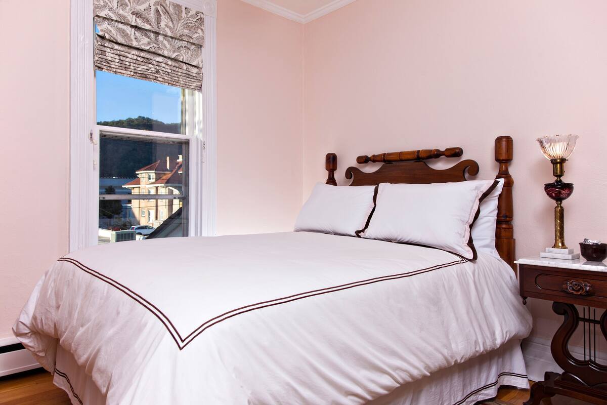 Una cama bien hecha con cabecera de madera, una mesita de noche con lámpara, y una ventana con vistas a edificios.
