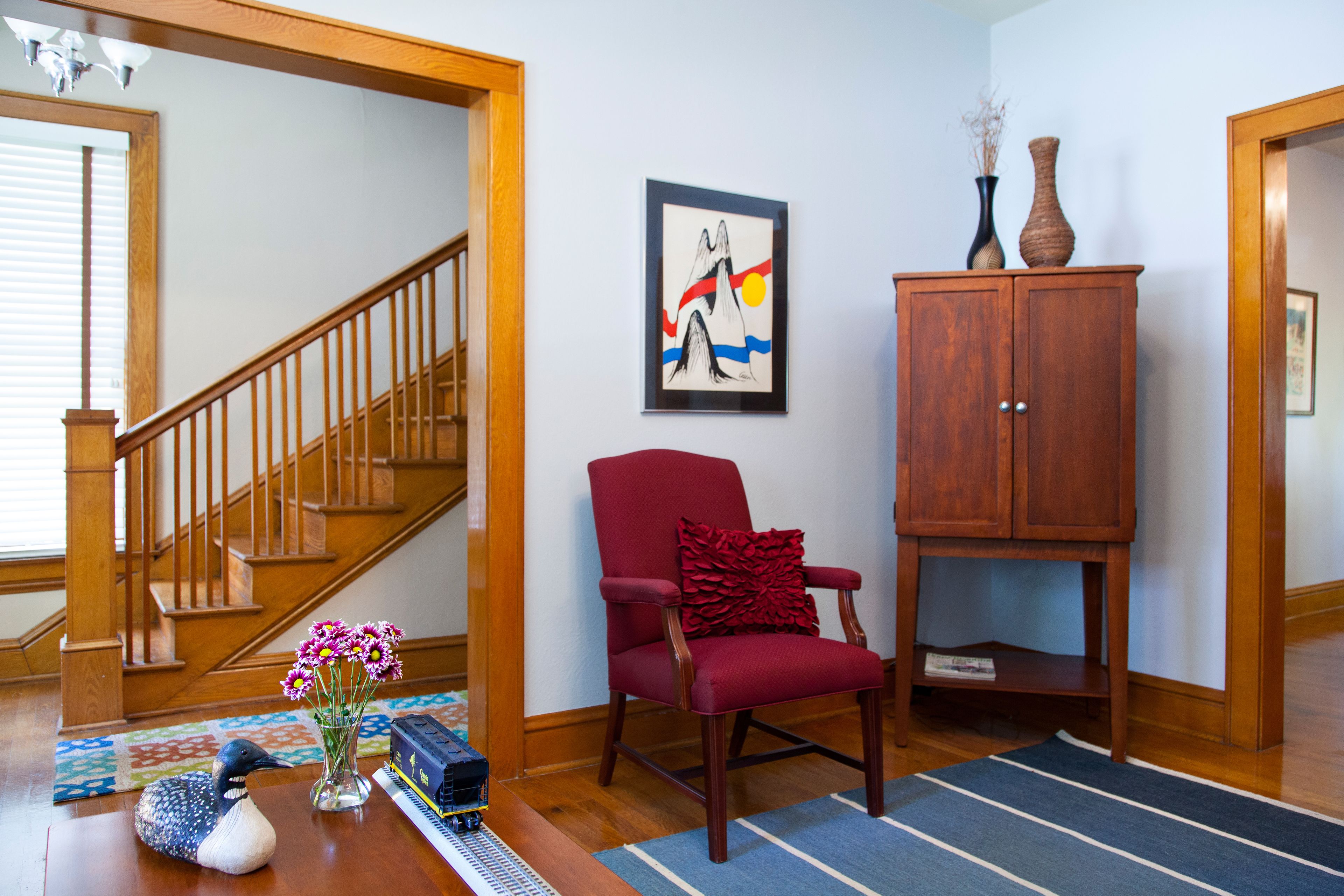 A living room with a wooden staircase, a red armchair, a wooden cabinet, and a framed artwork on the wall.