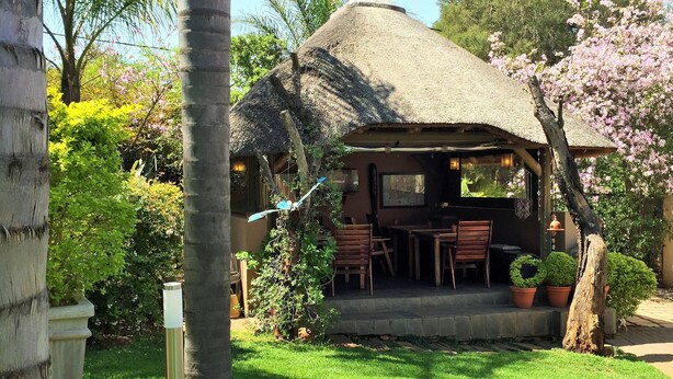 A thatched-roof structure with outdoor seating, surrounded by lush greenery and flowering plants. - Khayamanzi Guesthouse (Hartbeespoort)
