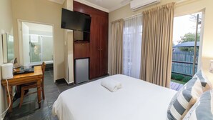 A hotel room with a bed, a desk with a chair, a TV mounted on the wall, a mini fridge, and a window with curtains. - Khayamanzi Guesthouse (Hartbeespoort)