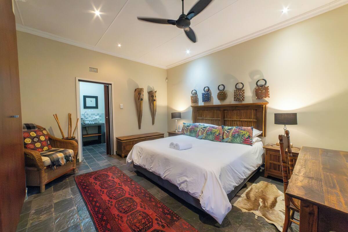 A spacious bedroom with a large bed, a wooden nightstand, a wooden table, a ceiling fan, and a red patterned rug.