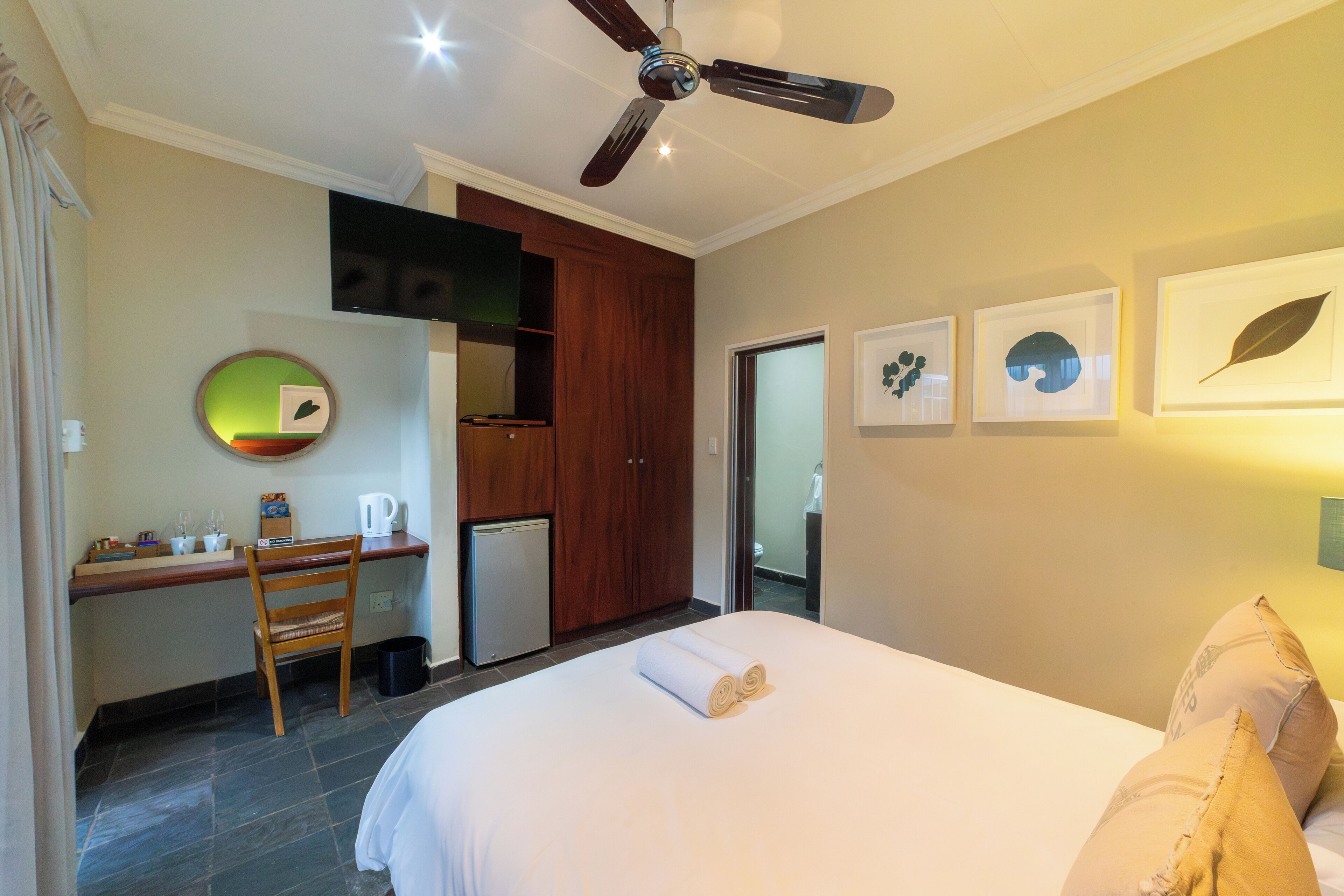 Une chambre d’hôtel équipée d’un lit, d’un bureau, d’une chaise, d’un ventilateur de plafond et d’œuvres d’art accrochées au mur.