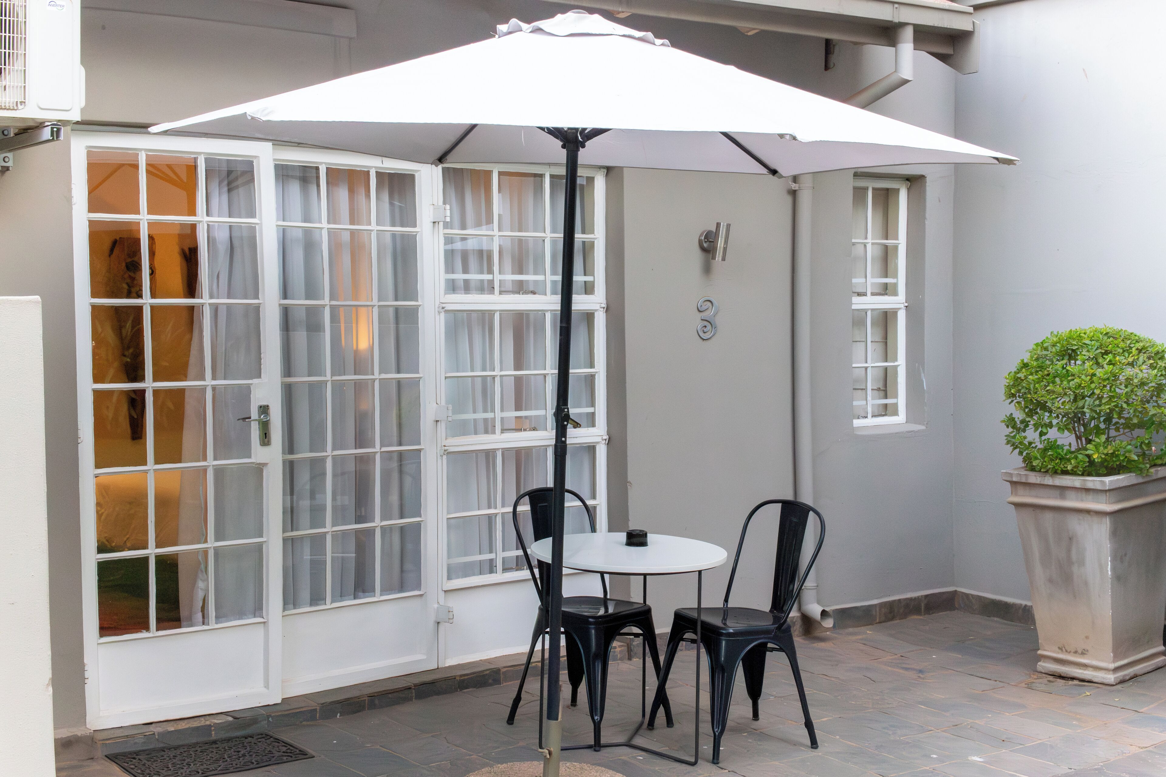 Een terras met een witte tafel en twee stoelen onder een grote parasol, een witte deur met ruitpanelen en een plant in een pot.