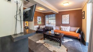 A living room with a sofa, a coffee table, a TV mounted on the wall, and a brick wall. - Khayamanzi Guesthouse (Hartbeespoort)