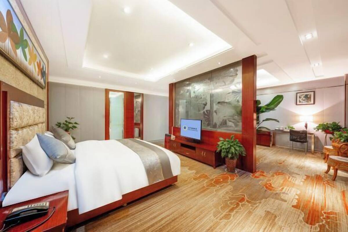 A hotel room with a bed, TV, desk, chair, and potted plants.