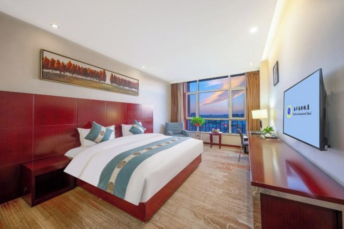 A hotel room with a large bed, a desk, a TV, and a window with a view of the sunset.