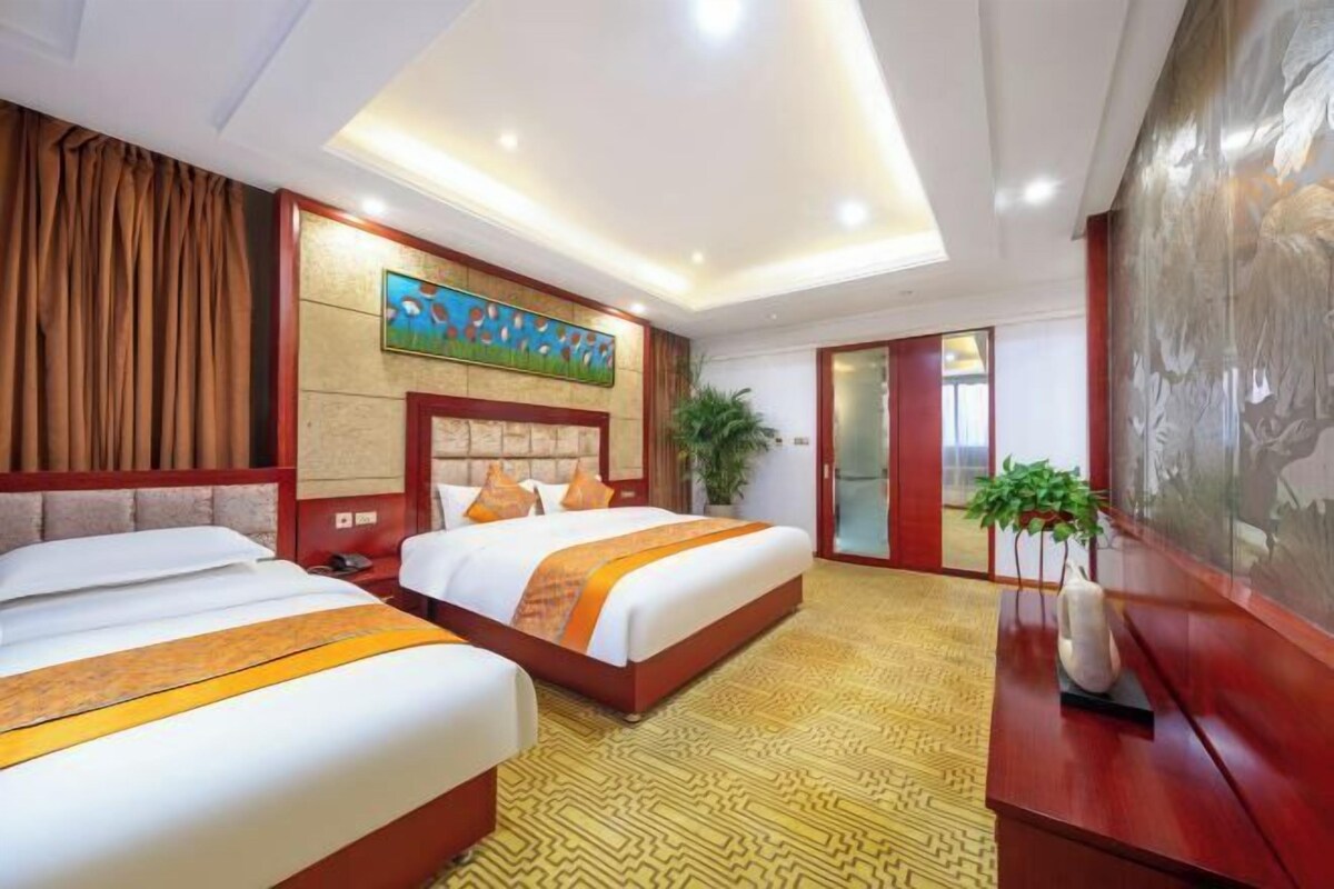 Habitación de hotel con dos camas, un cuadro grande en la pared, una planta en maceta y un jarrón decorativo sobre una mesa de madera.