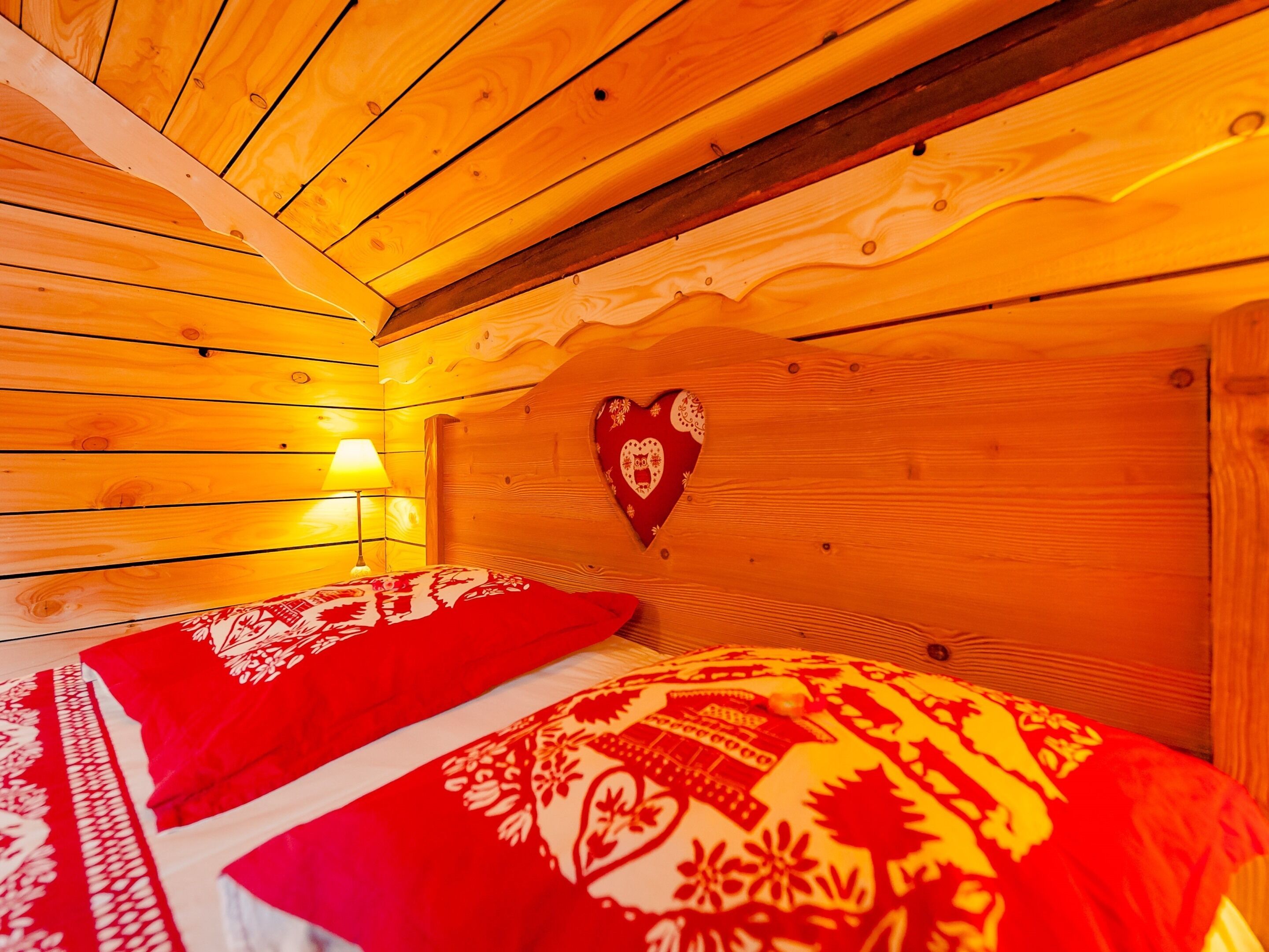 A wooden cabin room with a bed, a wall-mounted heart-shaped decoration, and a lamp.