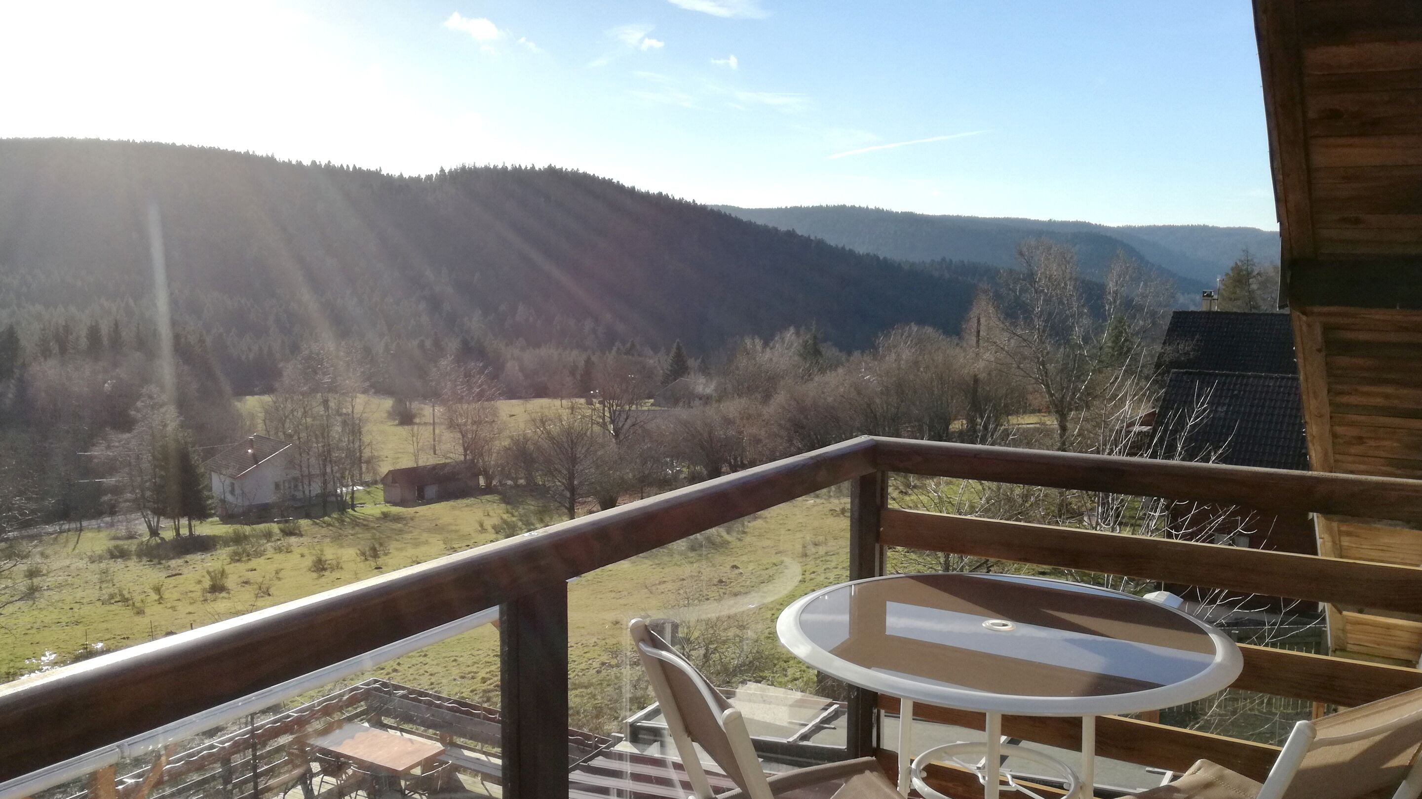A balcony with a table and chairs, overlooking a valley with mountains and a small building.