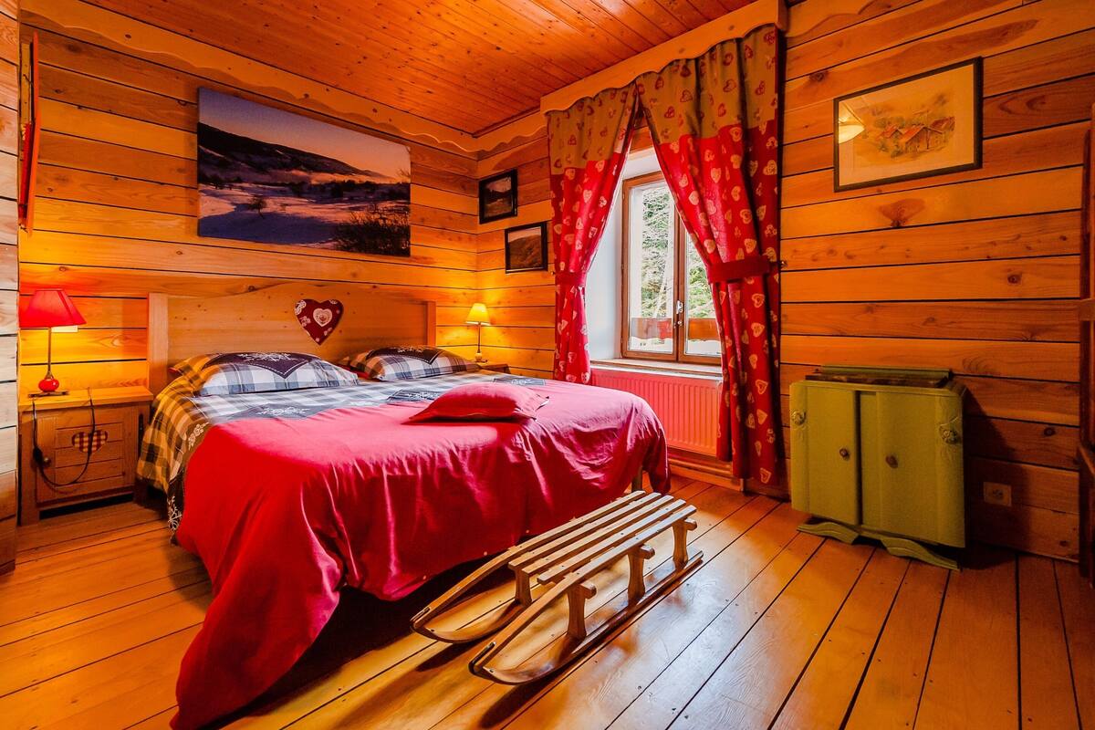 A cozy cabin-style bedroom with a wooden ceiling, a bed with red bedding, a bench, and a green cabinet.