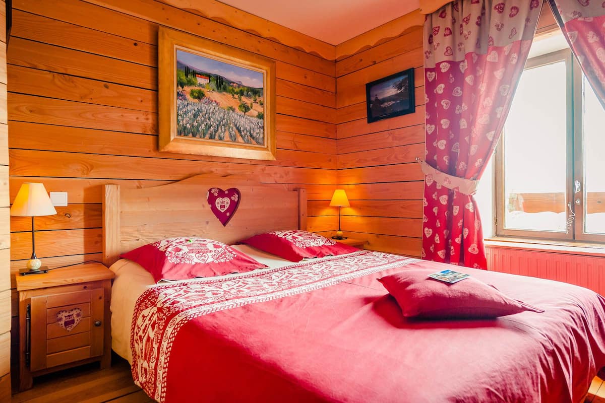 A wooden cabin-style bedroom with a bed, two bedside lamps, a heart-shaped decoration, and a window with curtains.