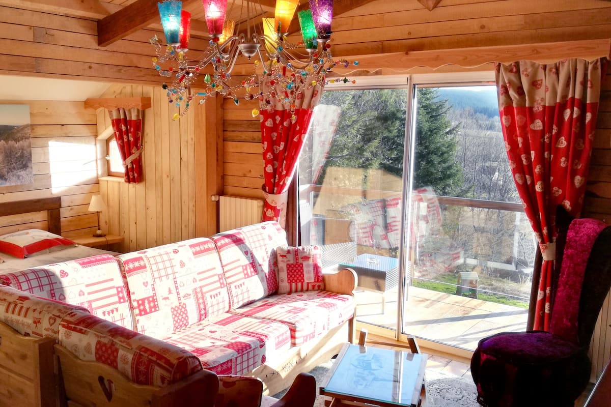 A cozy cabin interior with a wooden interior, a patterned sofa, a chandelier, and a large window with curtains.