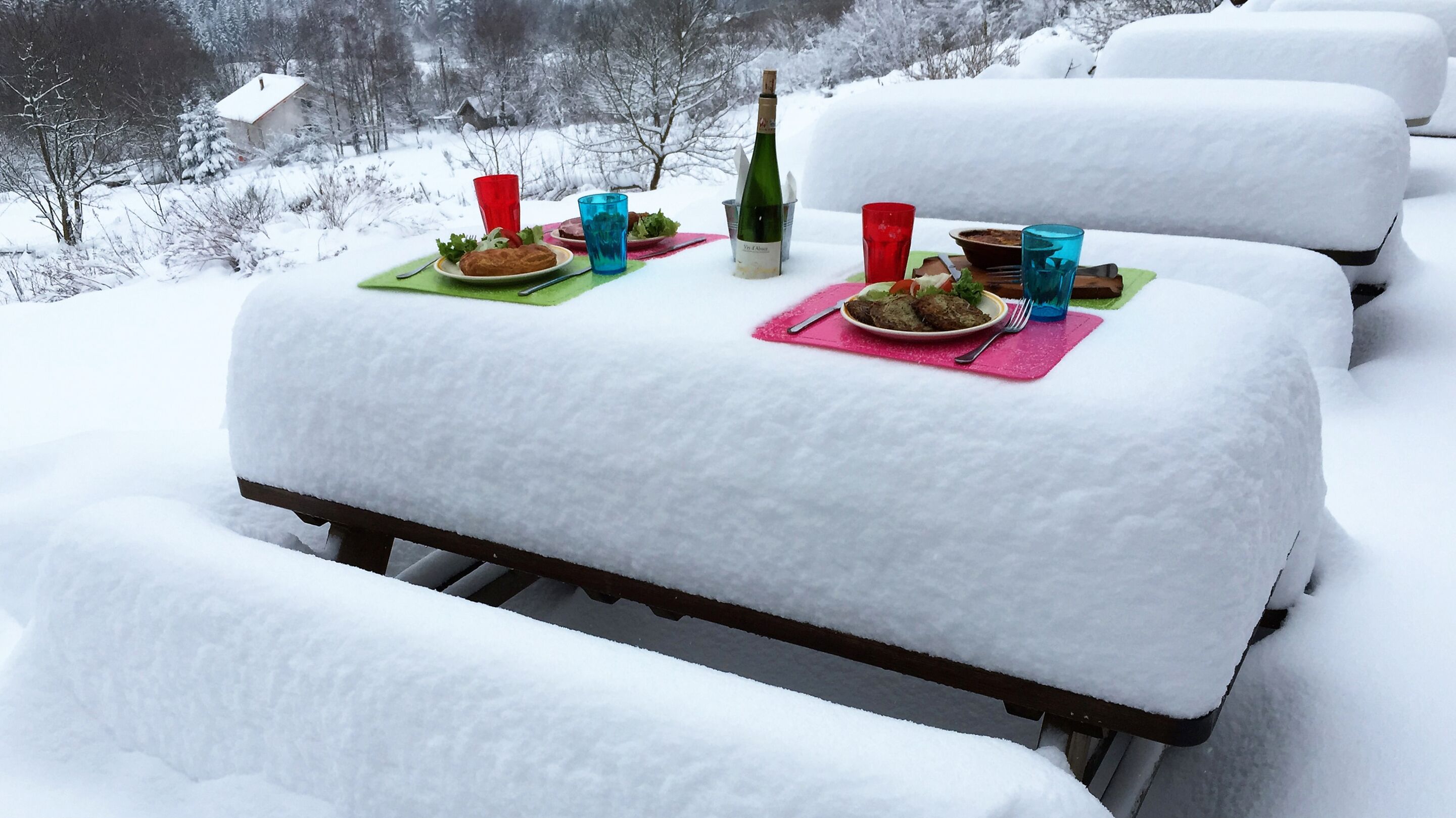 A snow-covered outdoor dining table with plates of food, glasses, and a bottle of wine.