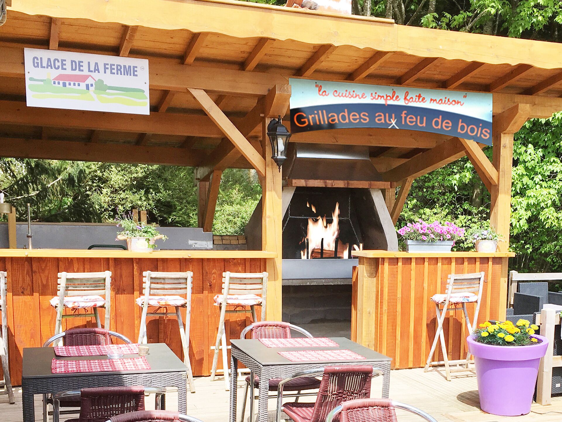 An outdoor dining area with wooden tables and chairs, a grill station, and signs indicating a restaurant and a simple home-style cuisine.