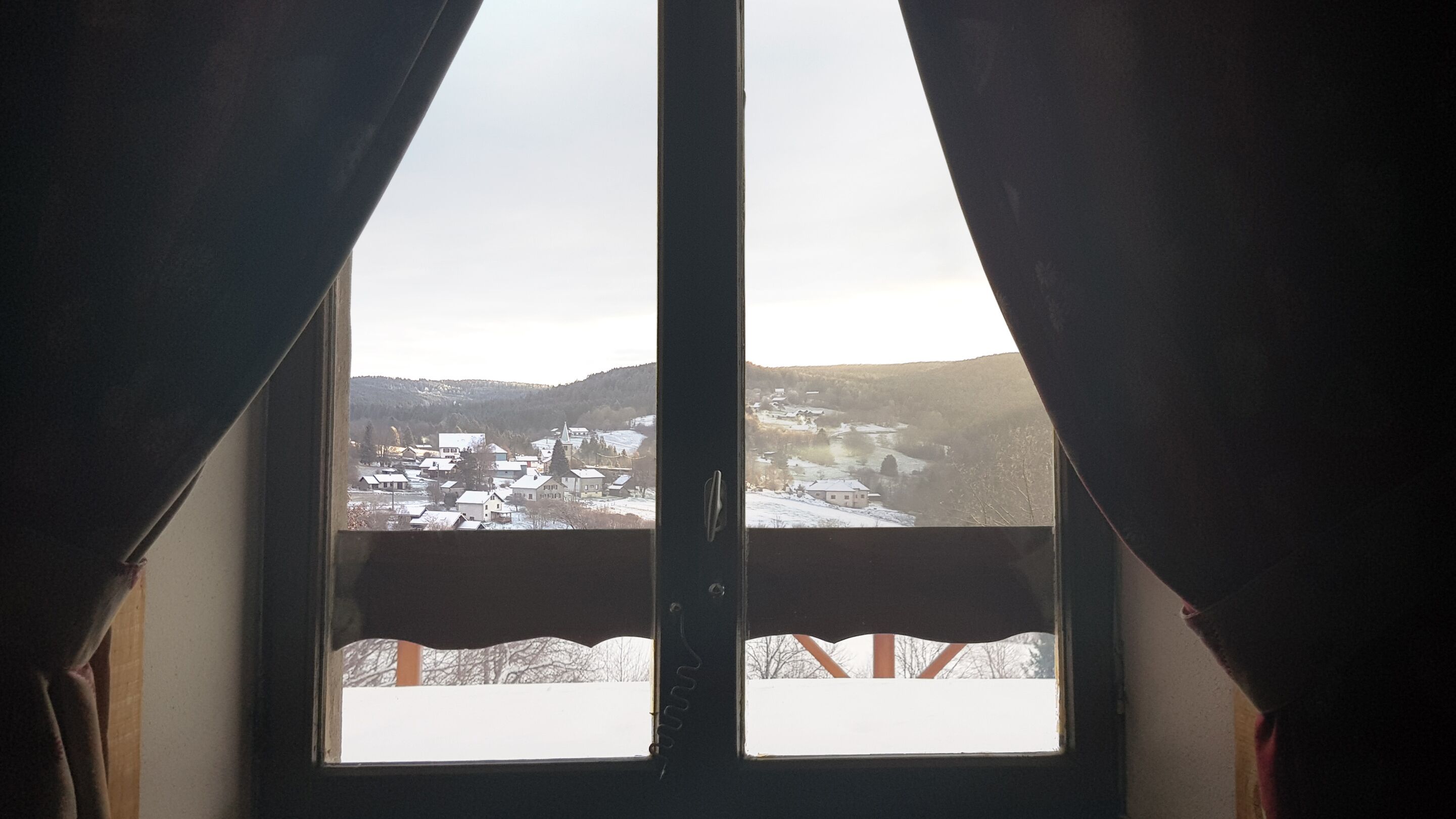Une fenêtre avec des rideaux, une vue sur un village enneigé et une montagne en arrière-plan.