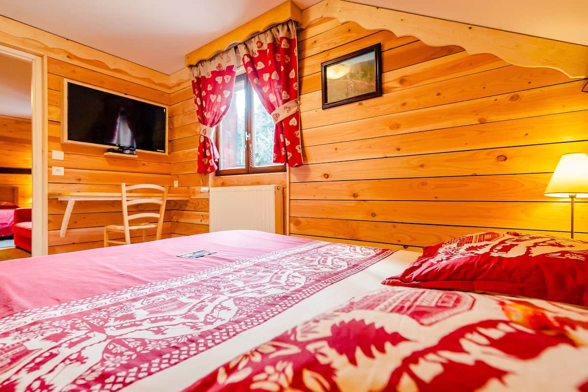 A wooden cabin room with a bed, a chair, a television, and a window with curtains.