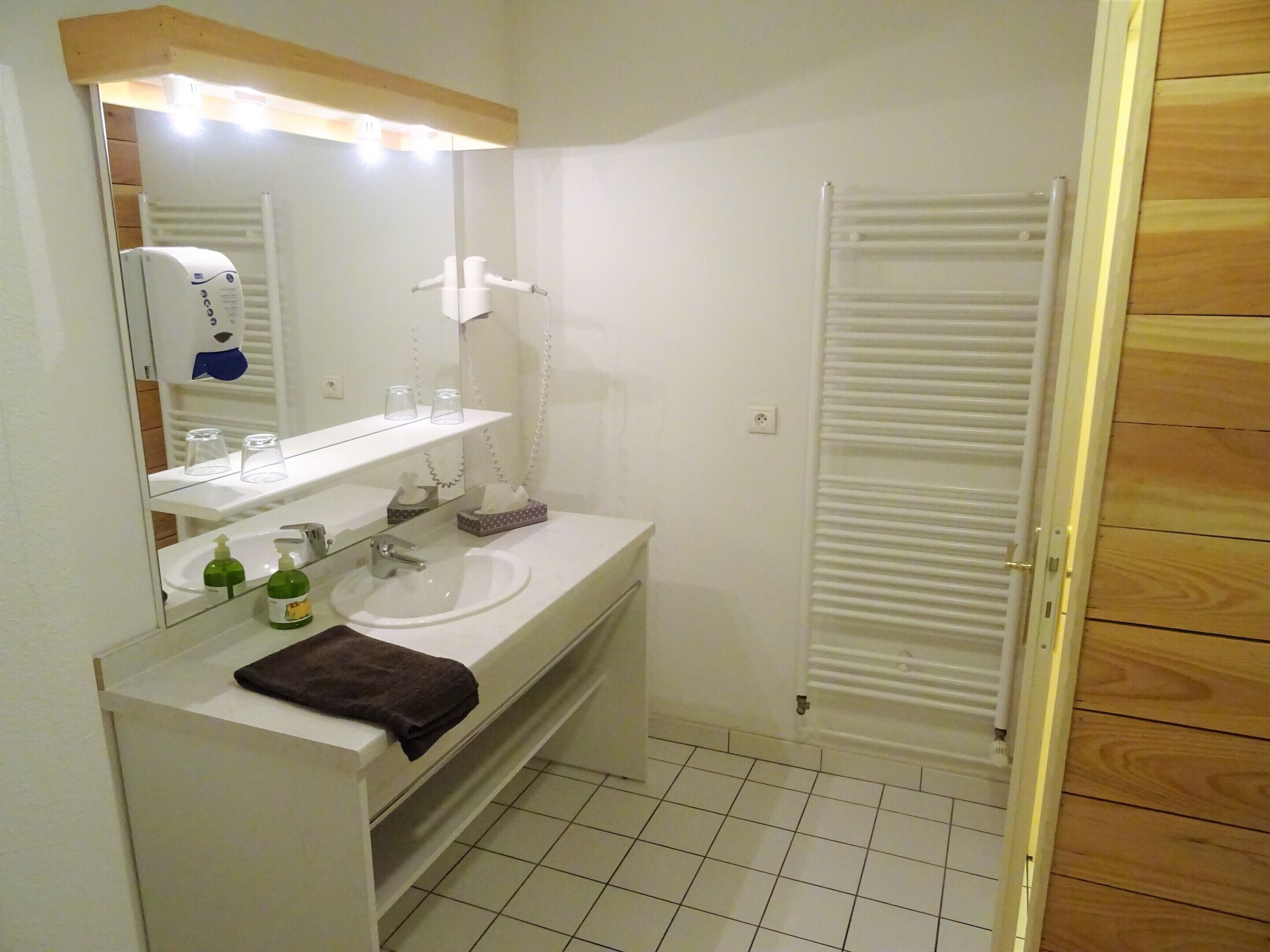 Casa de banho com lavatório branco, espelho e porta-toalhas. Um toalha está pendurada no porta-toalhas, e uma embalagem de sabonete está no lavatório.
