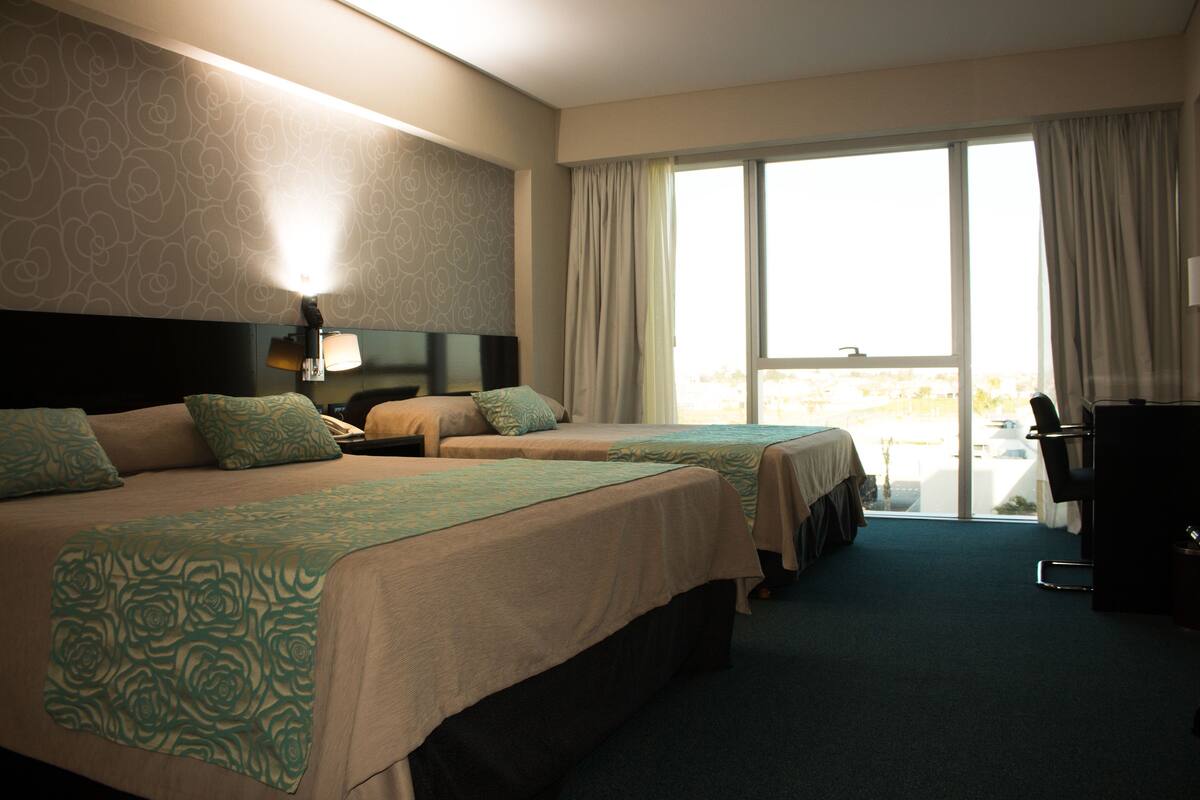 Une chambre d’hôtel avec deux lits, un bureau, une chaise et une grande fenêtre avec des rideaux.