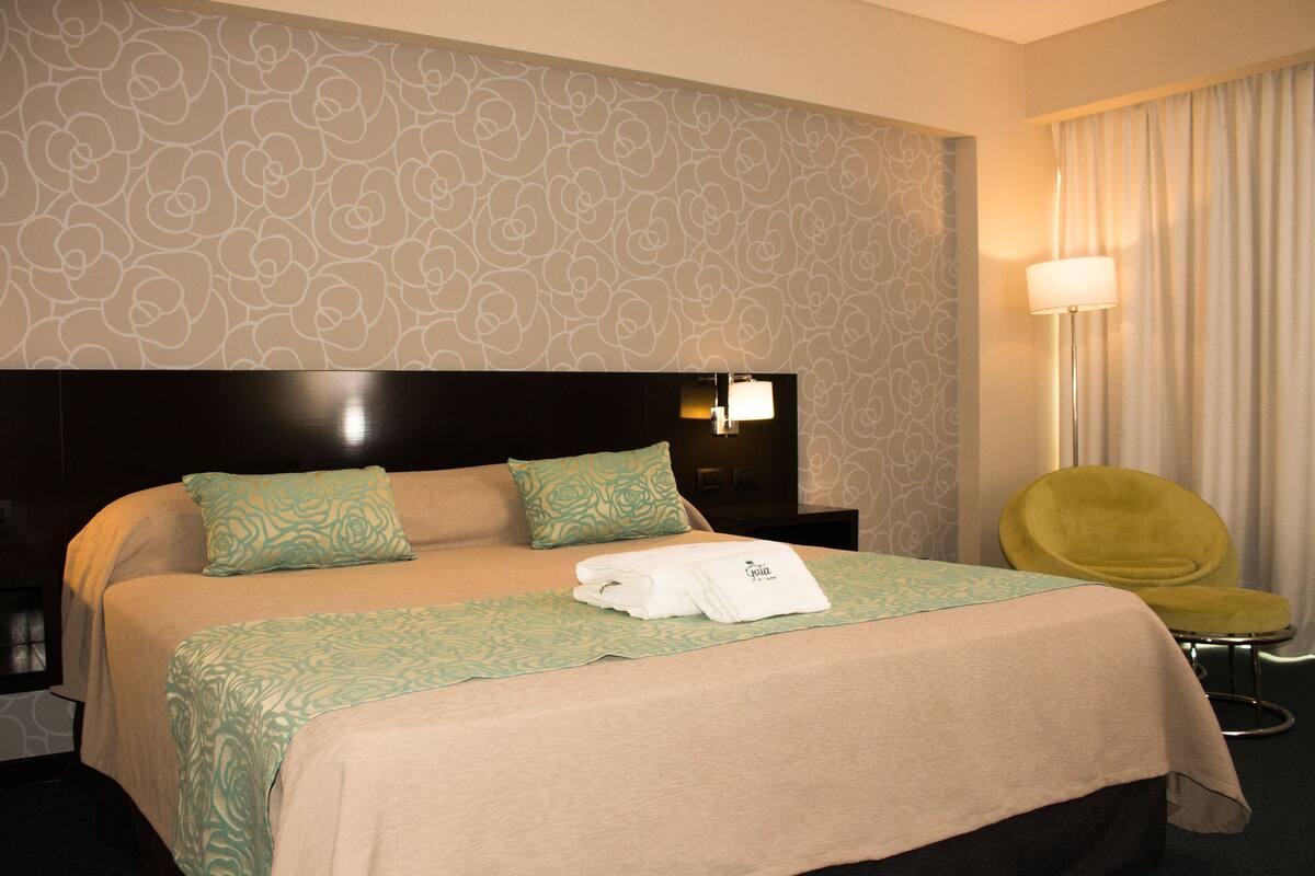 Une chambre d’hôtel avec un grand lit, une tête de lit, une table de chevet, une lampe, une chaise et une fenêtre avec des rideaux.