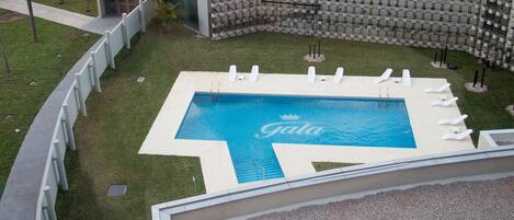 Uma área de piscina com o logotipo 'Gala', cercada por uma cerca branca, incluindo cadeiras de lounge e um pequeno edifício.
