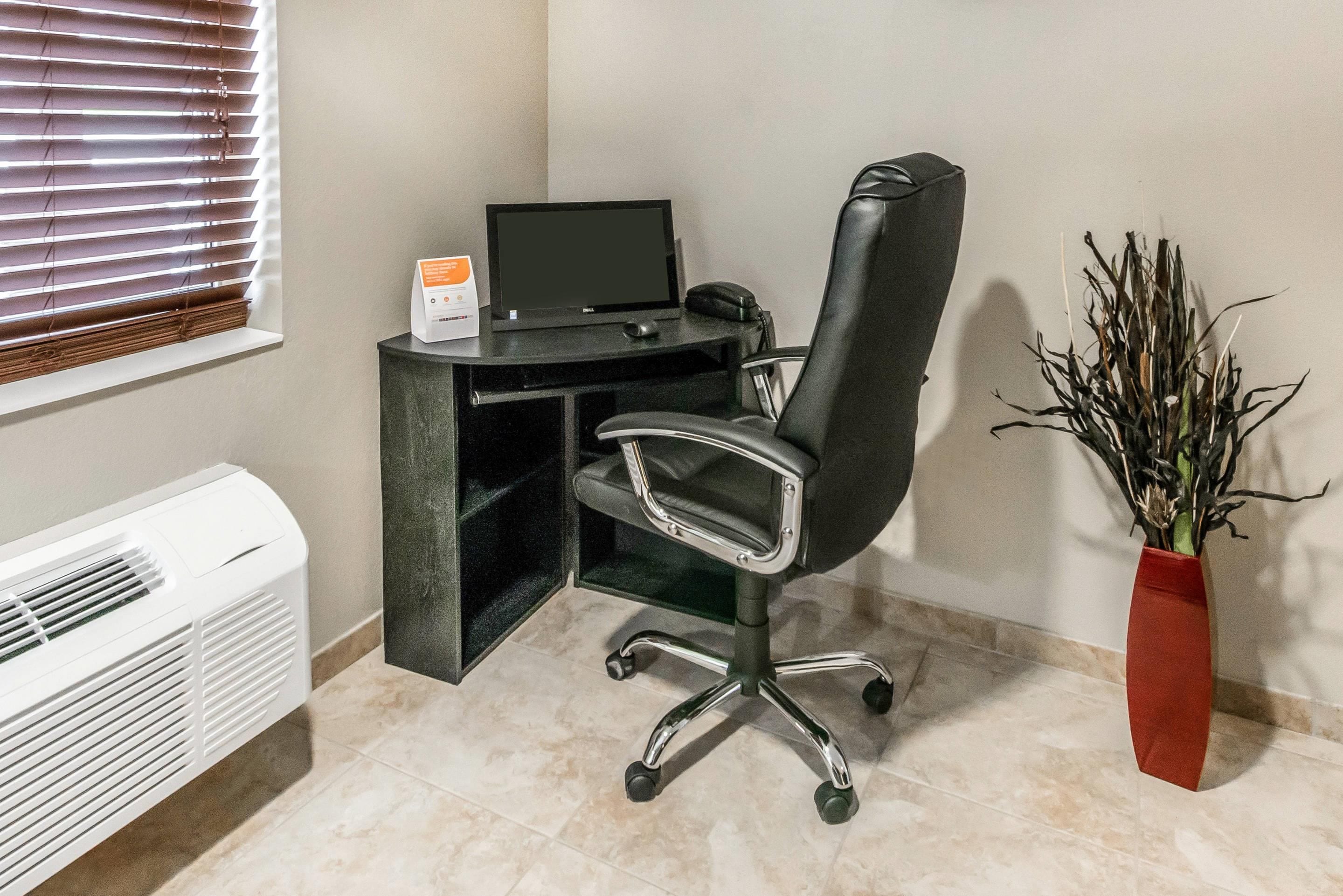 A corner office with a black desk, a computer, a chair, a radiator, and a potted plant.