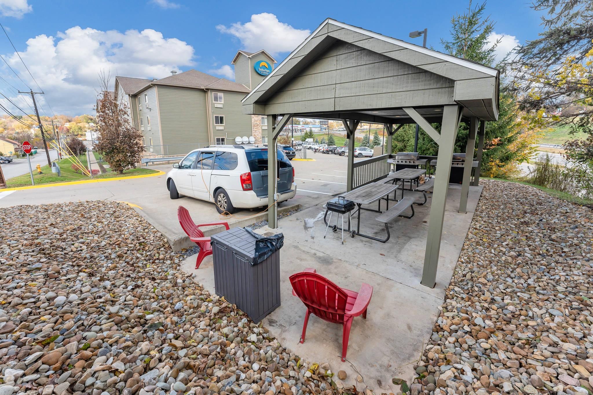 A covered outdoor area with picnic tables and a barbecue grill, surrounded by a gravel area and a parked vehicle.