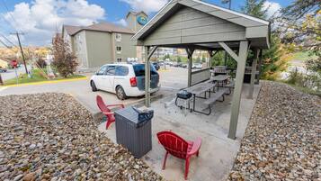 A covered outdoor area with picnic tables and a barbecue grill, surrounded by a gravel area and a parked vehicle.