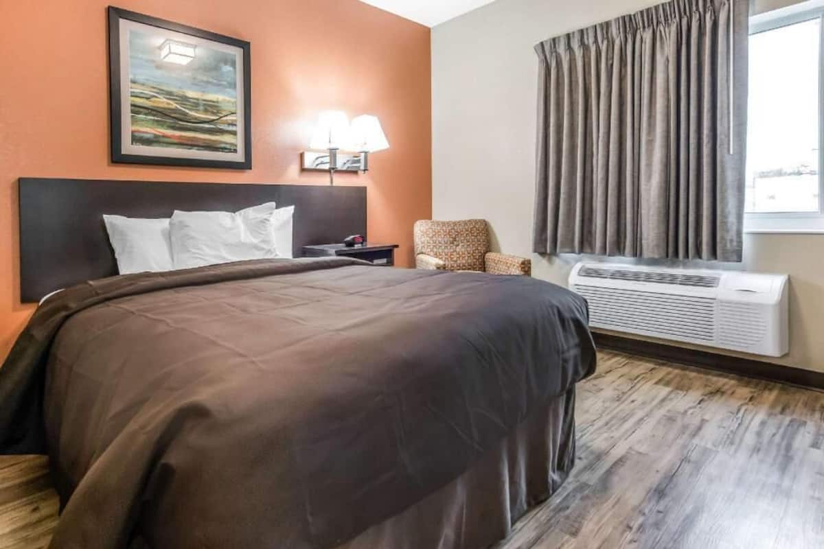 A hotel room with a large bed, a chair, a nightstand, a lamp, a window with curtains, and an air conditioning unit.