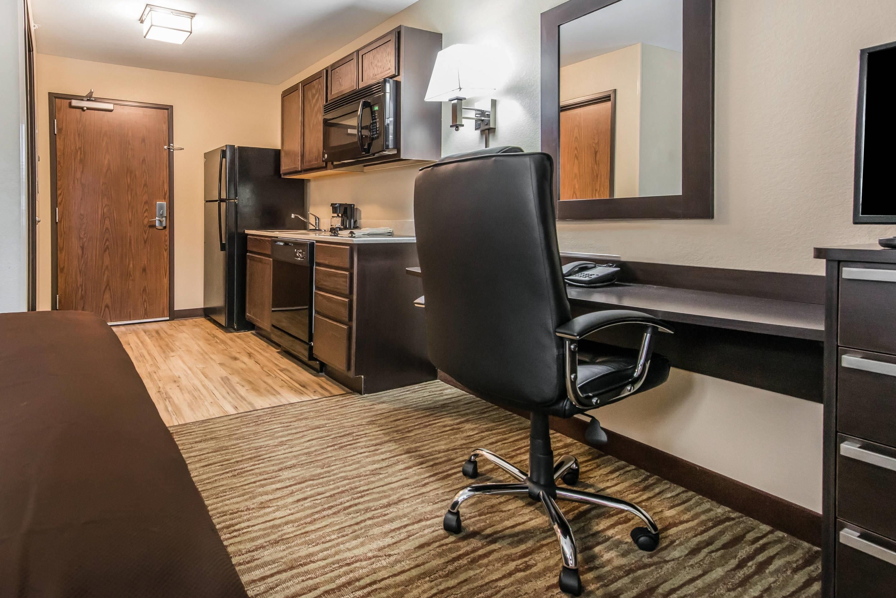 A hotel room with a kitchenette, a dining table, a black office chair, a desk with a computer, and a large mirror.