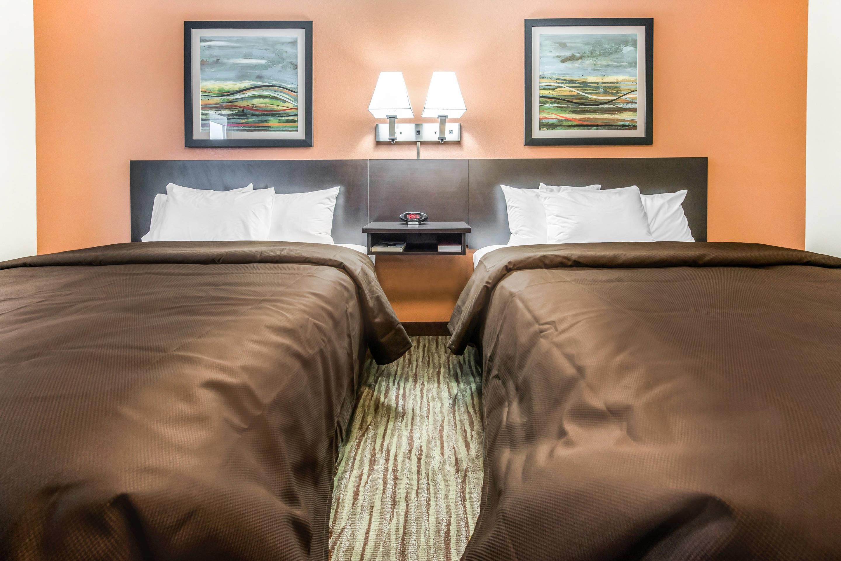 A hotel room with two beds, a nightstand, two wall-mounted lamps, and two framed artworks above the beds.