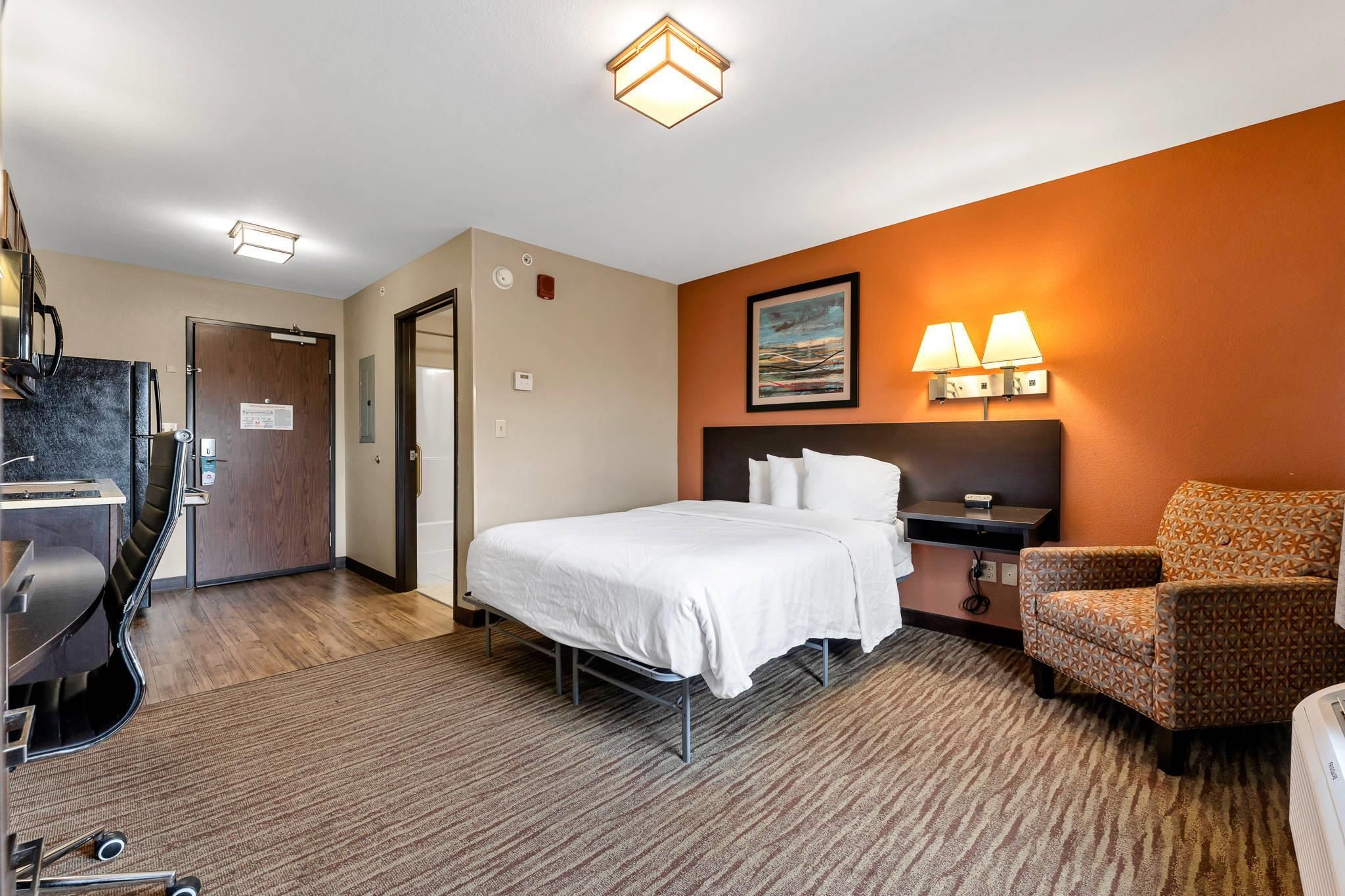 A hotel room with a bed, a chair, a desk, and a refrigerator.