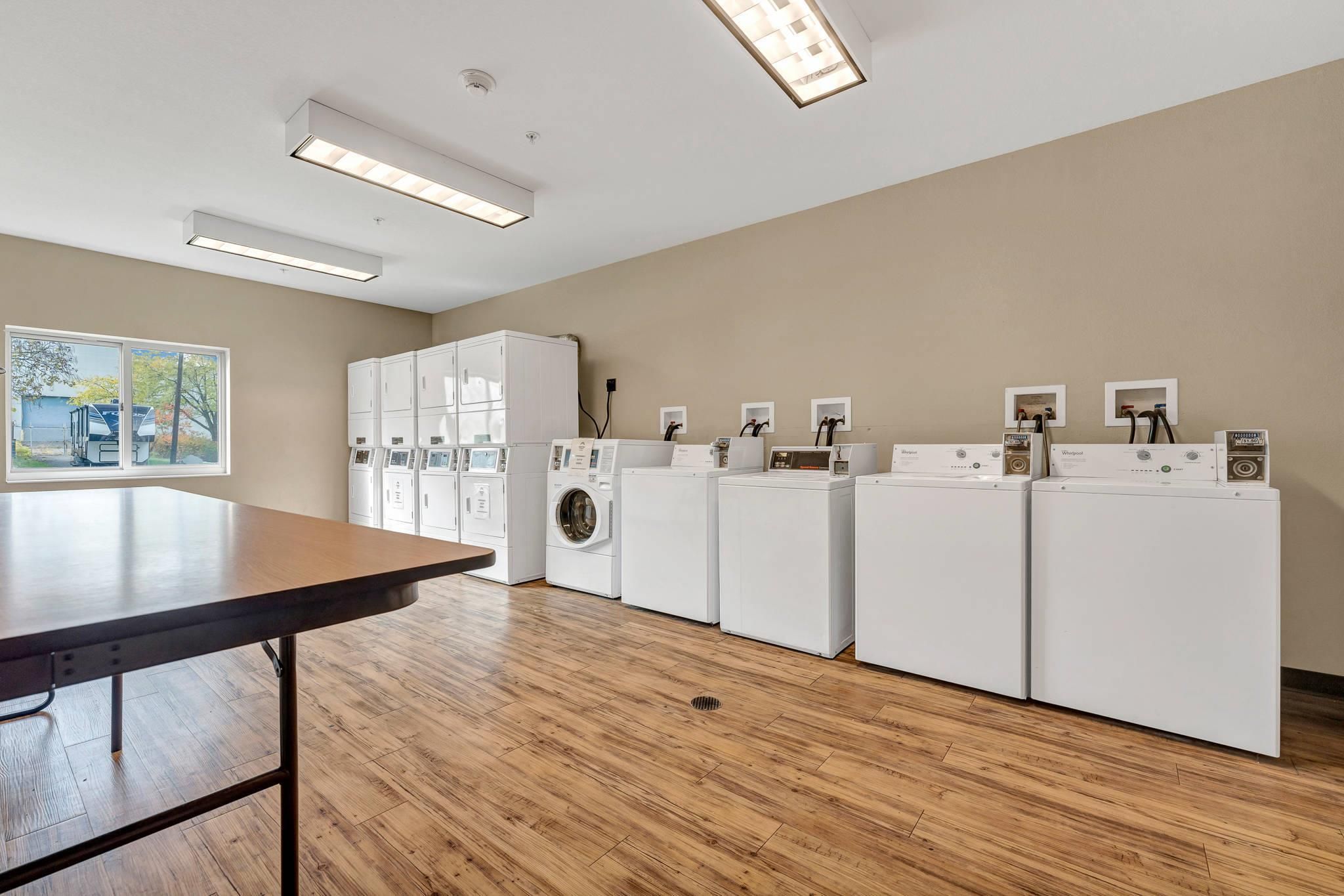 A laundry room with multiple washing machines, a table, and a window with a view of a vehicle and trees.