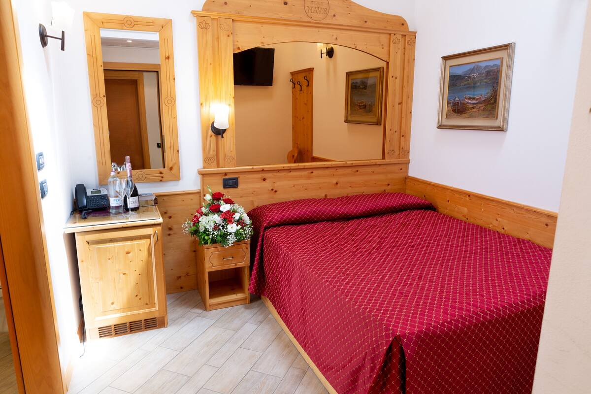 A wooden-framed bedroom with a bed, a wooden cabinet, a mirror, a wall-mounted TV, and a framed painting.